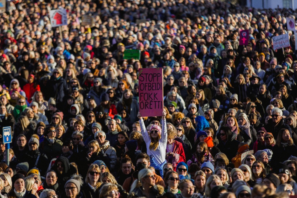 An audience of women and non-binary people with signs. A person with long red hair, sunglasses and a pink hat, in a white shirt, holds a pink sign lettered with black letters, that says "stop violence, fuck injustice". 